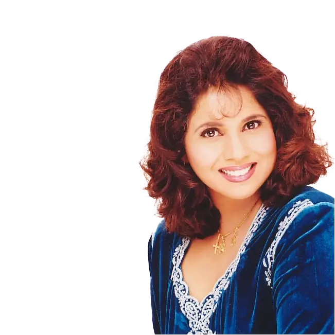 Hema Sardesai, smiling and wearing a blue velvet outfit with a necklace, against a black background.