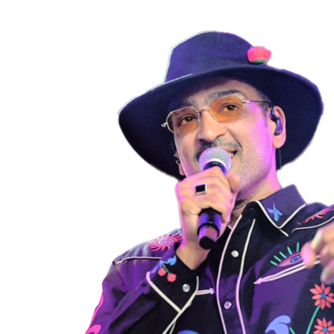 Ali Sethi wearing a black hat with a pink flower, glasses, and a patterned black shirt, holding a microphone